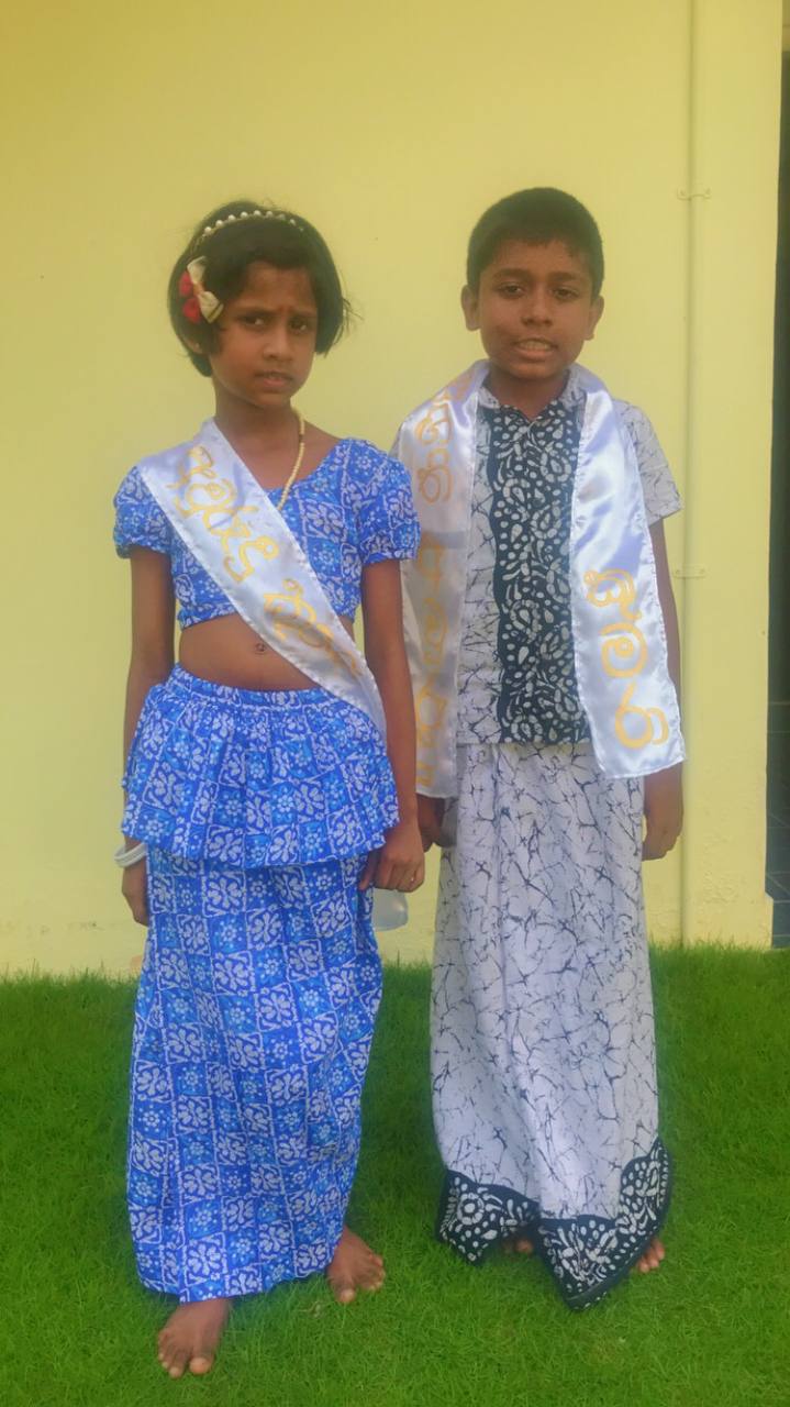 Children in vibrant traditional attire, barefoot on grass, posed against a light-yellow wall.