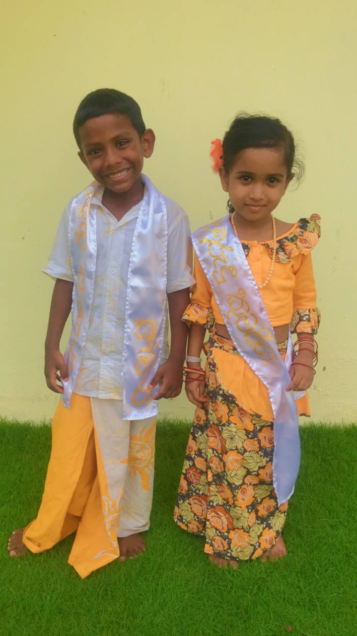 Two children in traditional attire smiling on grass against a plain wall.