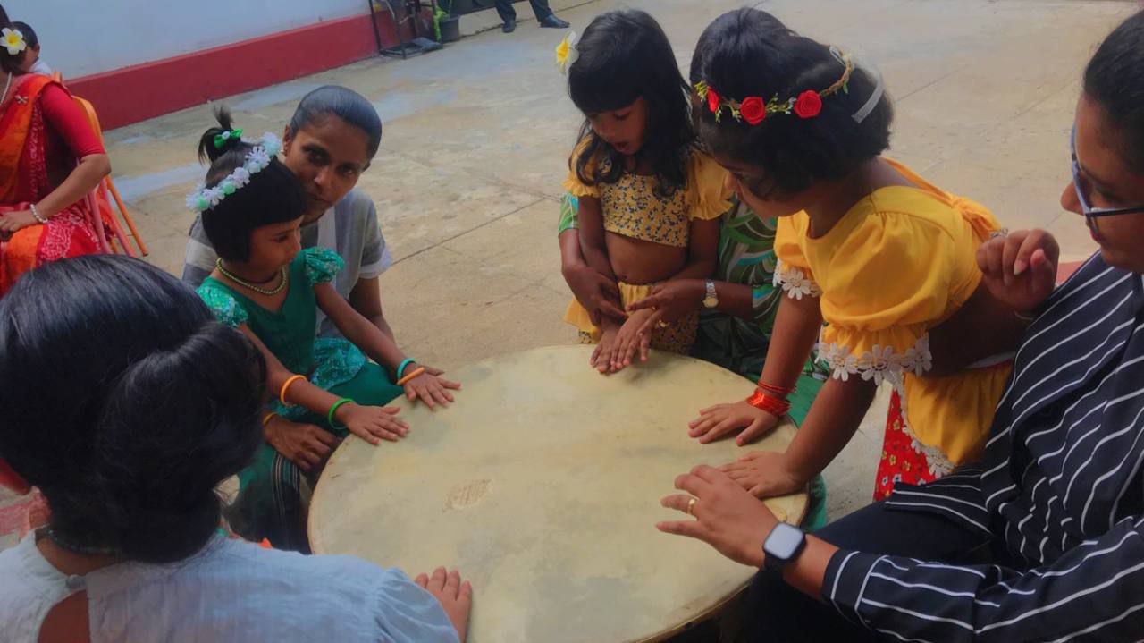 Children and adults playing a large communal drum in a festive outdoor setting.