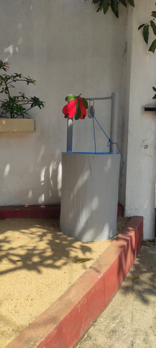Quaint garden corner with a grey well and red bucket, green foliage, and sandy ground.