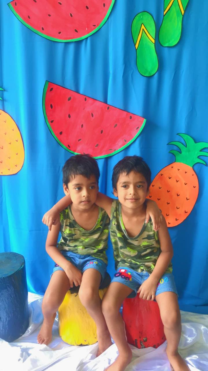 Twin boys in camo shirts on colorful stools with tropical fruit backdrop.