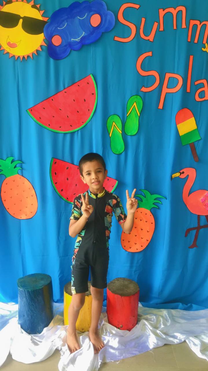 Boy in swimsuit poses with peace signs in front of colorful Summer Splash backdrop.