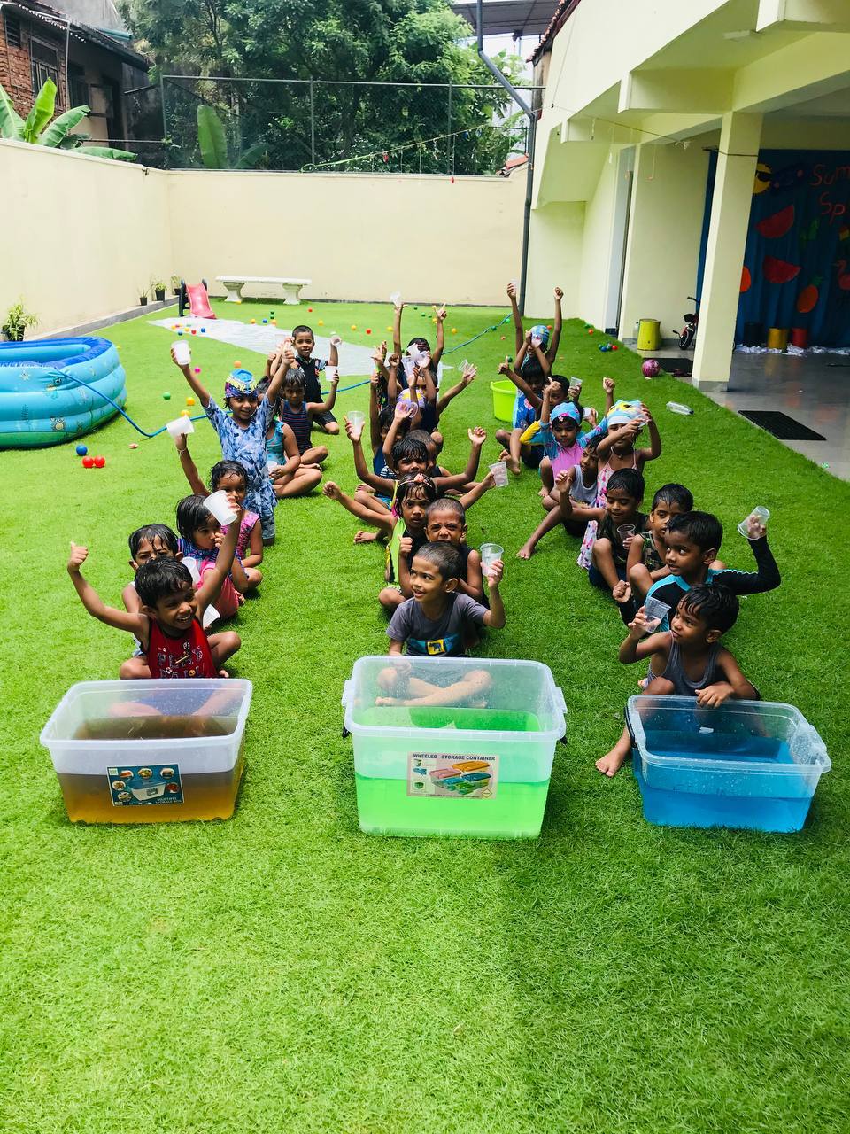 Children enjoying water play with colorful containers on a sunny artificial grass lawn.