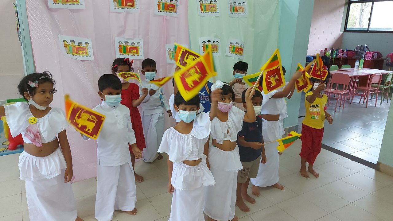 Children in festive attire wave Sri Lankan flags in a decorated classroom.