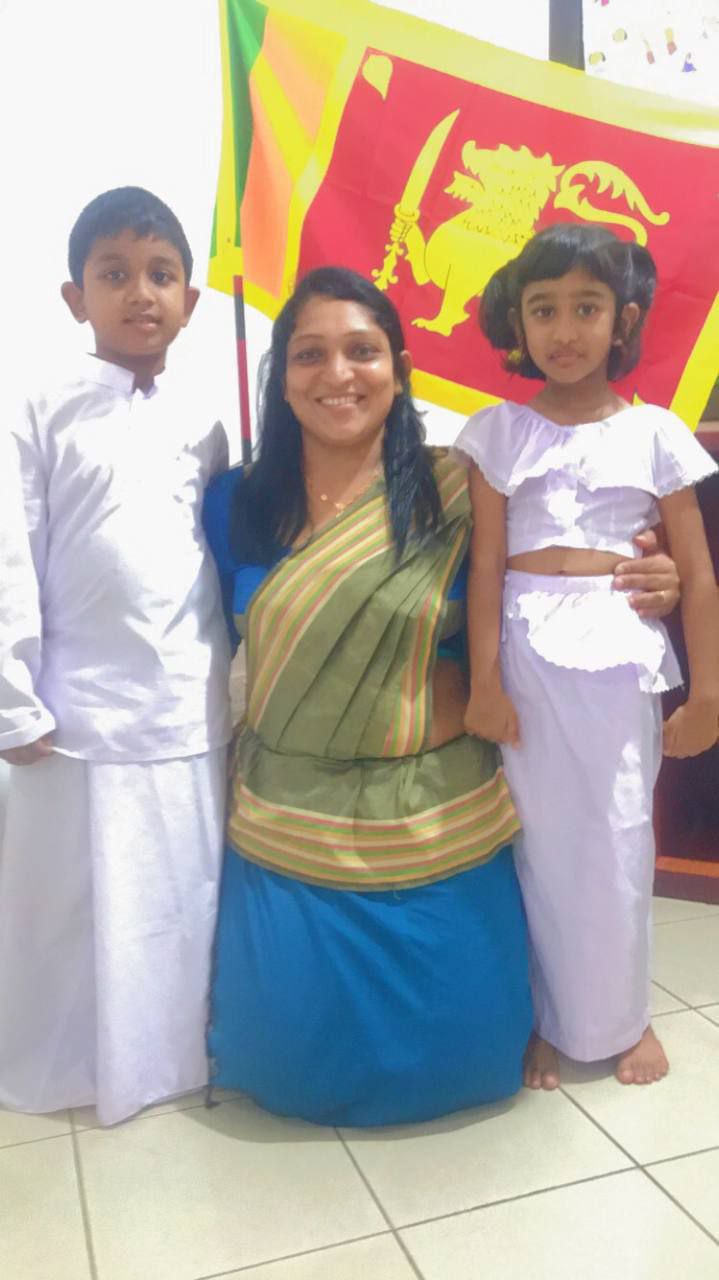 A woman and two children in traditional attire stand in front of a Sri Lankan flag.