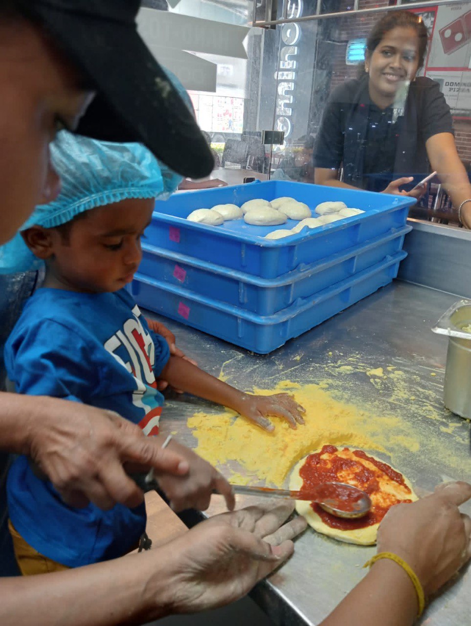 Child making pizza with adult guidance in a professional kitchen.