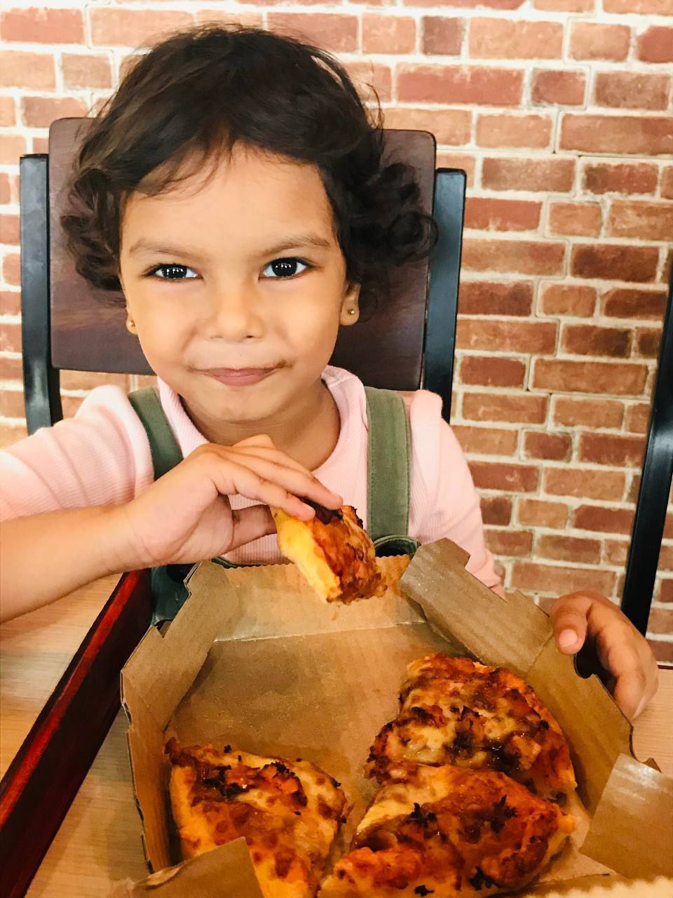 Child happily enjoying pizza in a cozy café with a brick wall background.