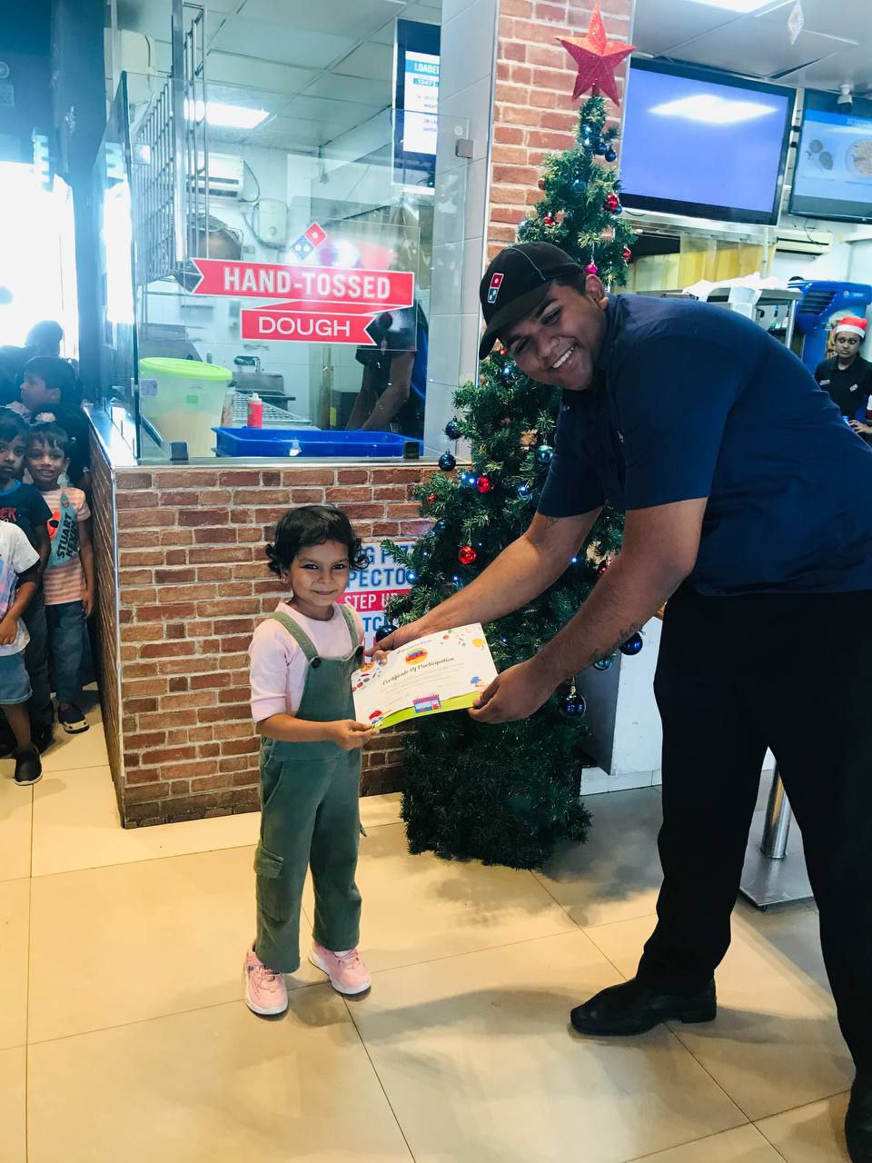 Child receives gift from employee at festive pizza restaurant with Christmas tree in background.