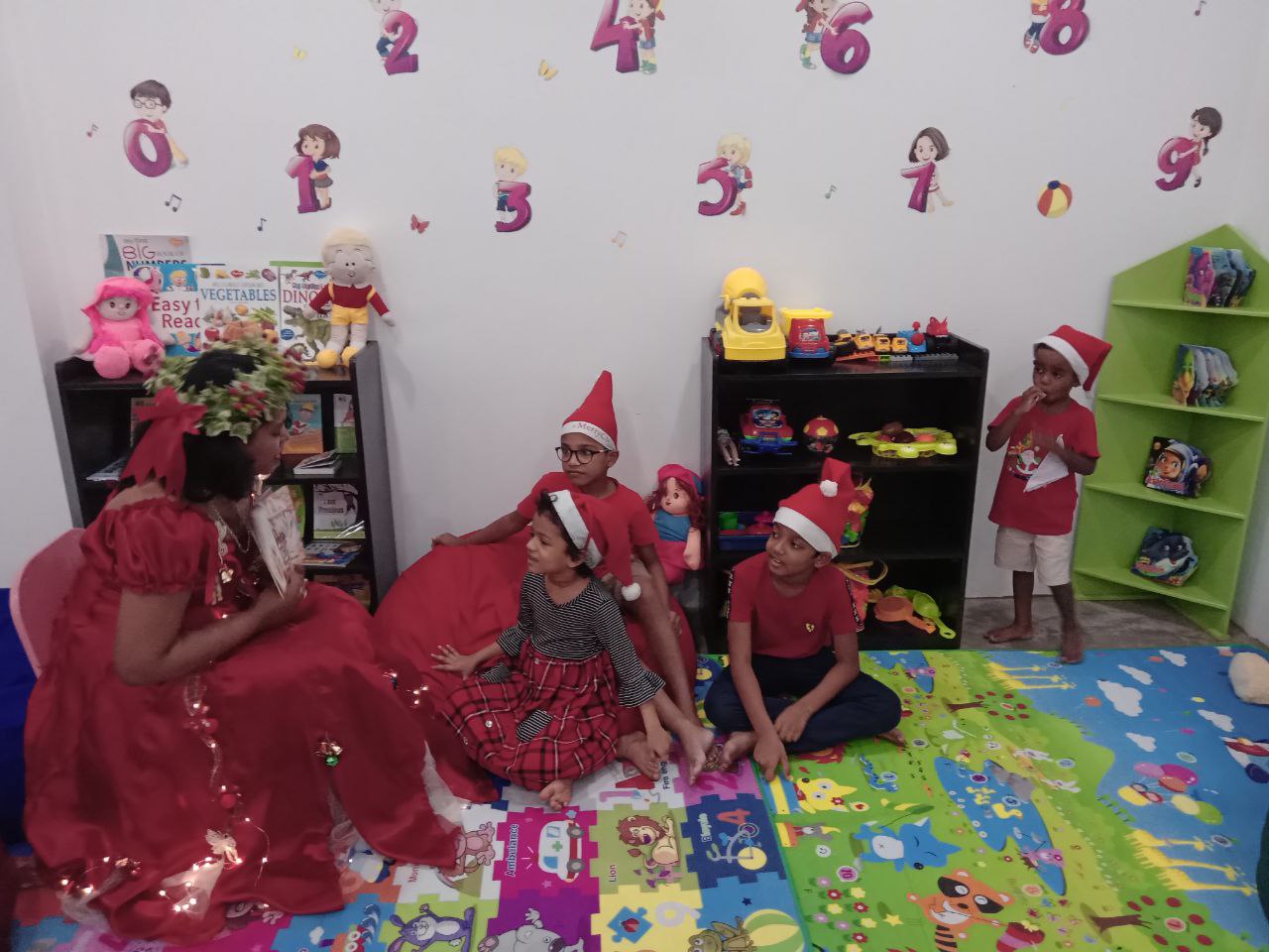 Festive playroom with children in Santa hats gathered around a woman reading a story.