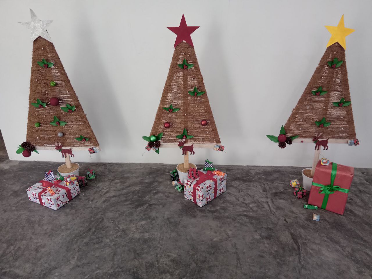 Handcrafted cardboard Christmas trees with stars, baubles, and gifts on a gray surface.