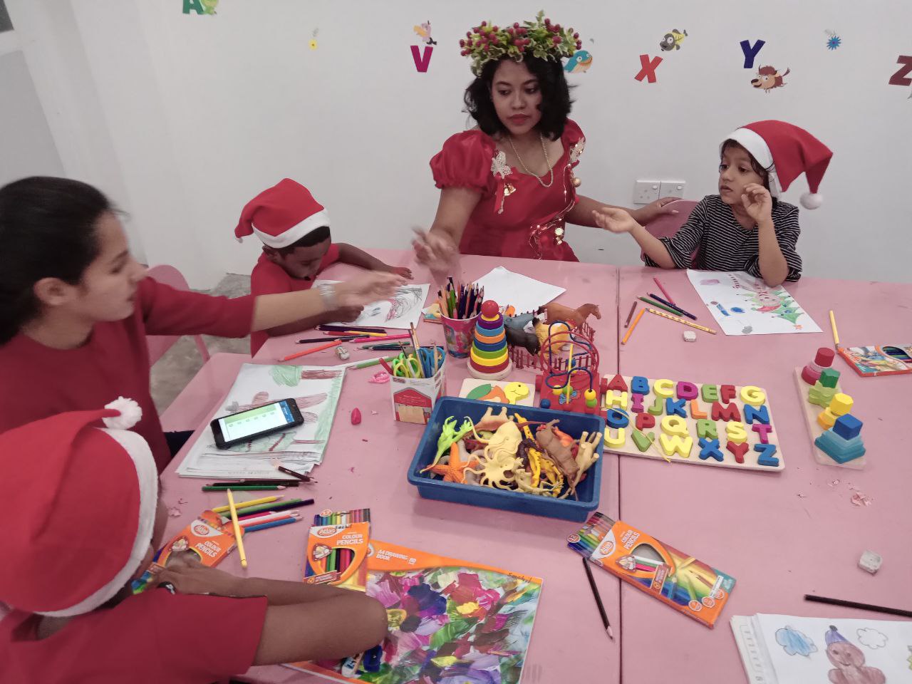 Kids and teacher in festive attire doing holiday crafts with art supplies at a colorful table.