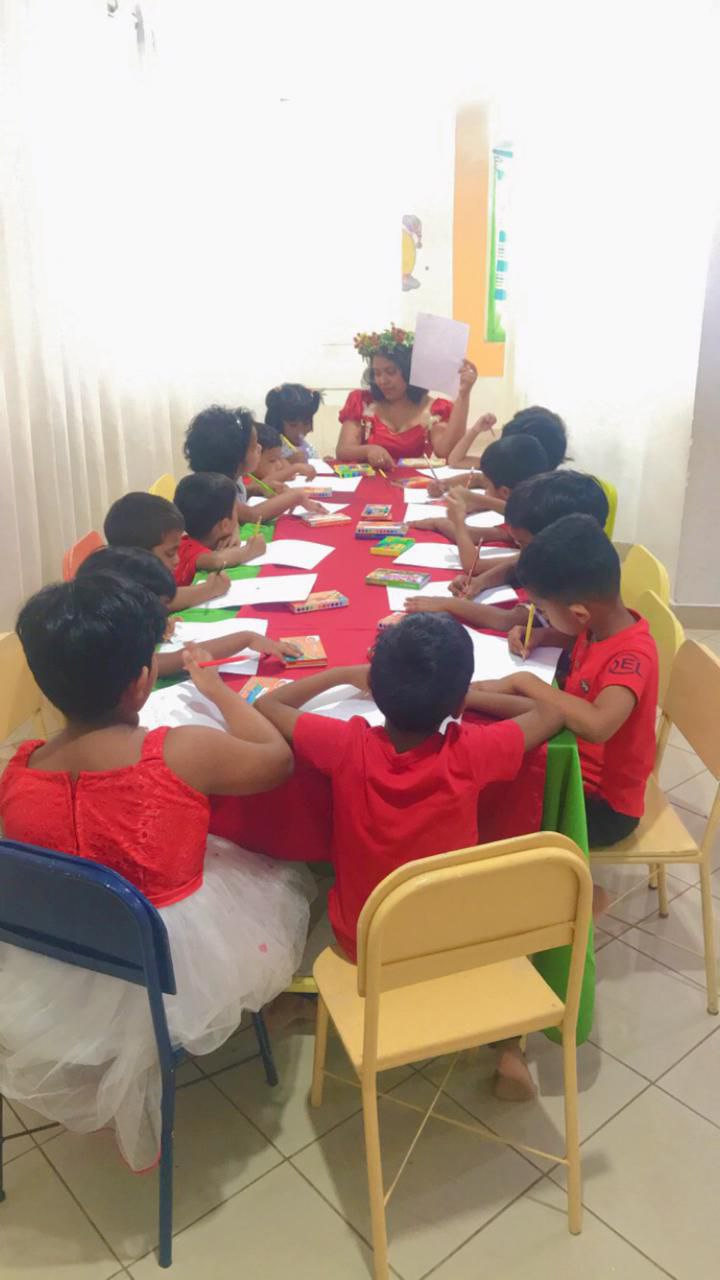 Children in festive outfits drawing at a table, guided by an adult in a flower crown.