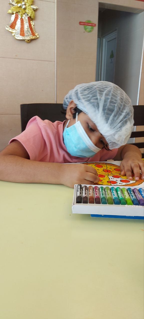 Child coloring with crayons, wearing a mask and hair net, in a festive indoor setting.