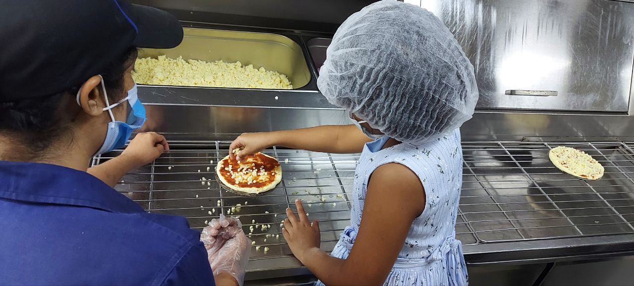 Child and adult making pizza in a professional kitchen, both wearing hairnets and masks.