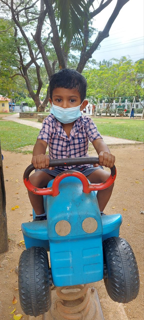 Child wearing mask rides blue toy car in sunny park.