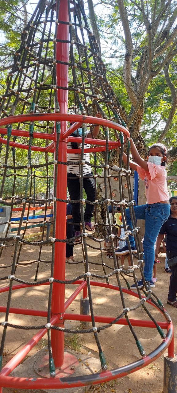 Children climbing large conical rope structure in tree-shaded playground.