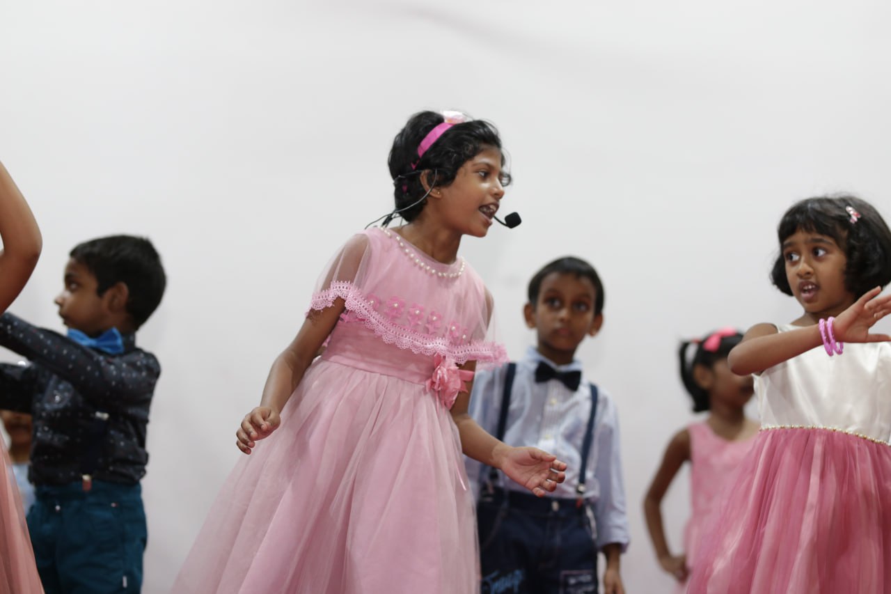Children performing on stage, girl in pink dress sings with headset, others dance in the background.