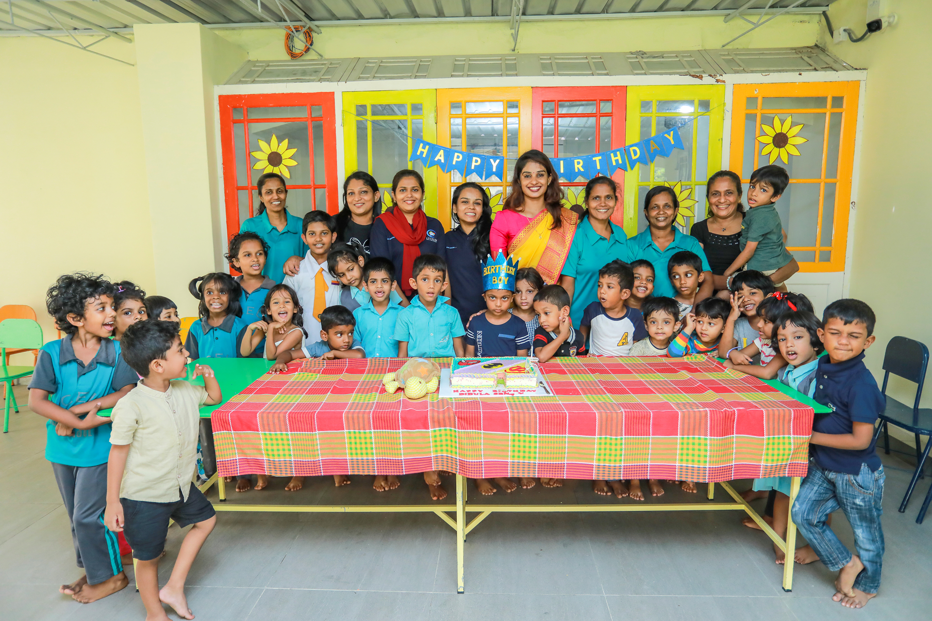Colorful community birthday party with children and adults around a decorated cake.