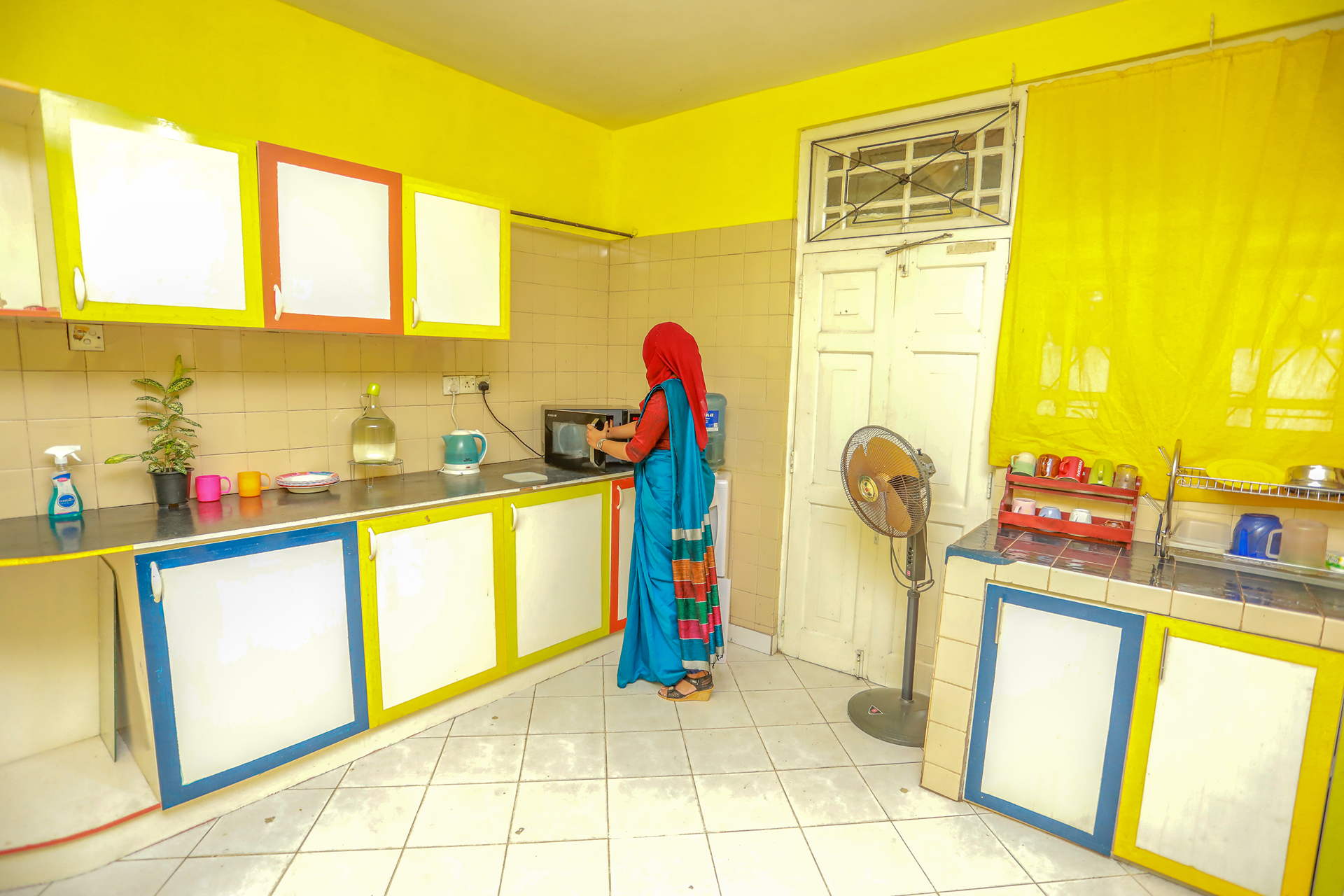 Person in traditional outfit using microwave in vibrant yellow kitchen with multicolored cabinets.