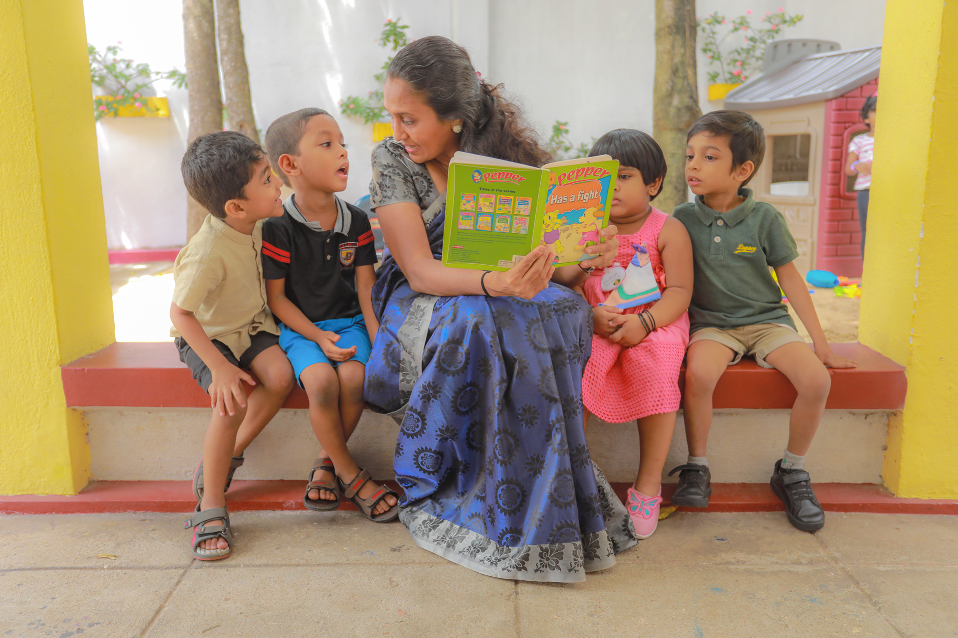 Woman reading to children in a colorful outdoor preschool setting.