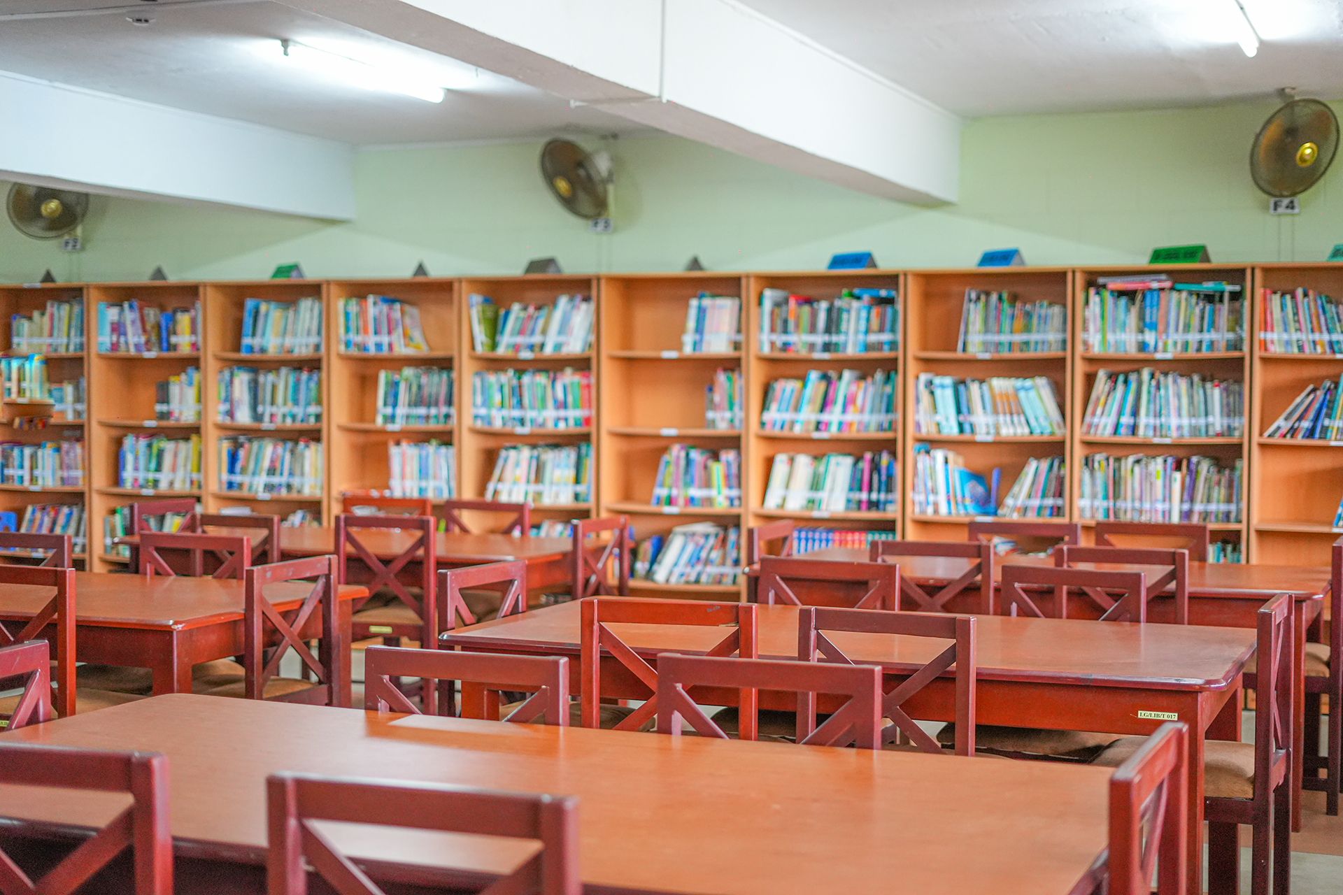 Spacious library with wooden tables, chairs, bookshelves, and ceiling fans.