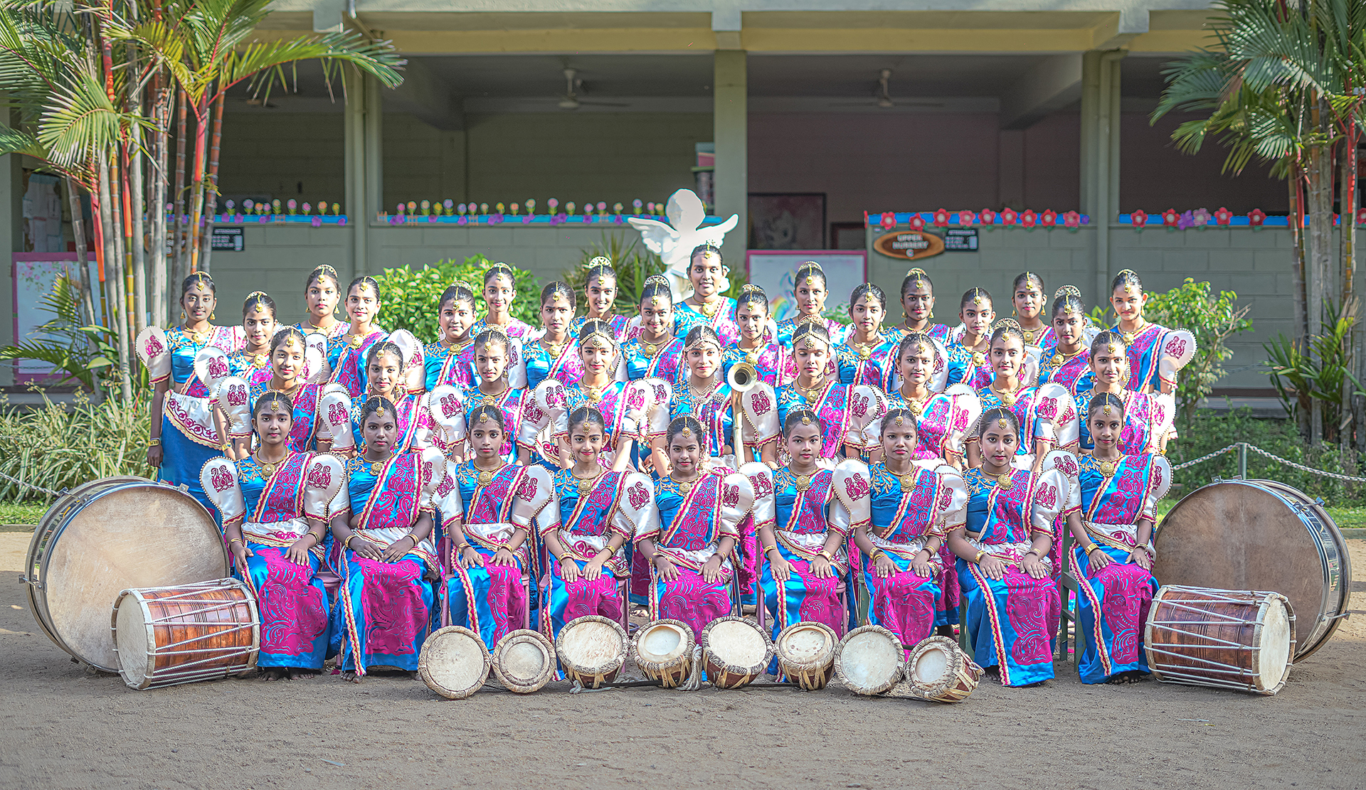 Girls in traditional attire with drums in front of a school building.