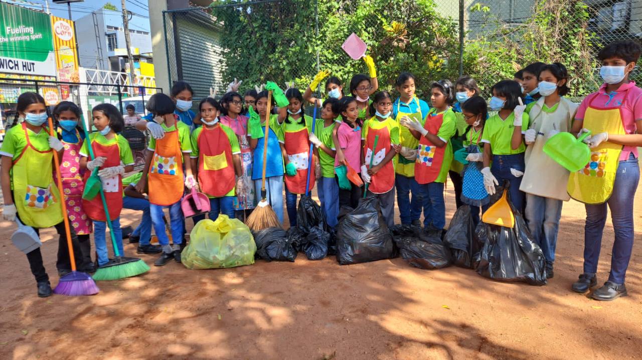 Children in colorful aprons and masks, cleaning up an urban outdoor area with brooms and trash bags.