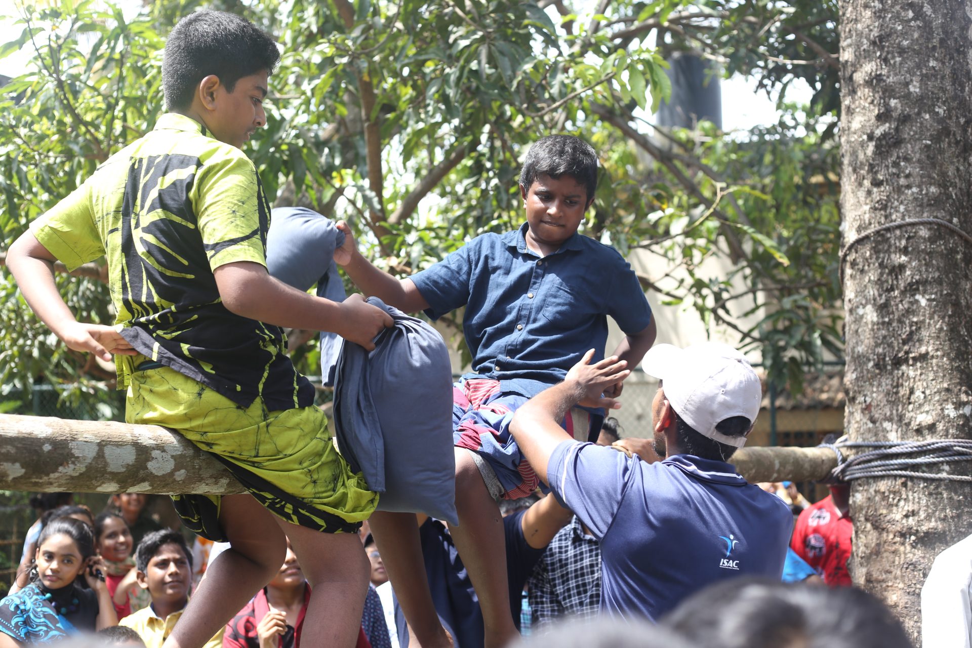 Boys on beam pillow fight, supervised by an adult, crowd watches in park.