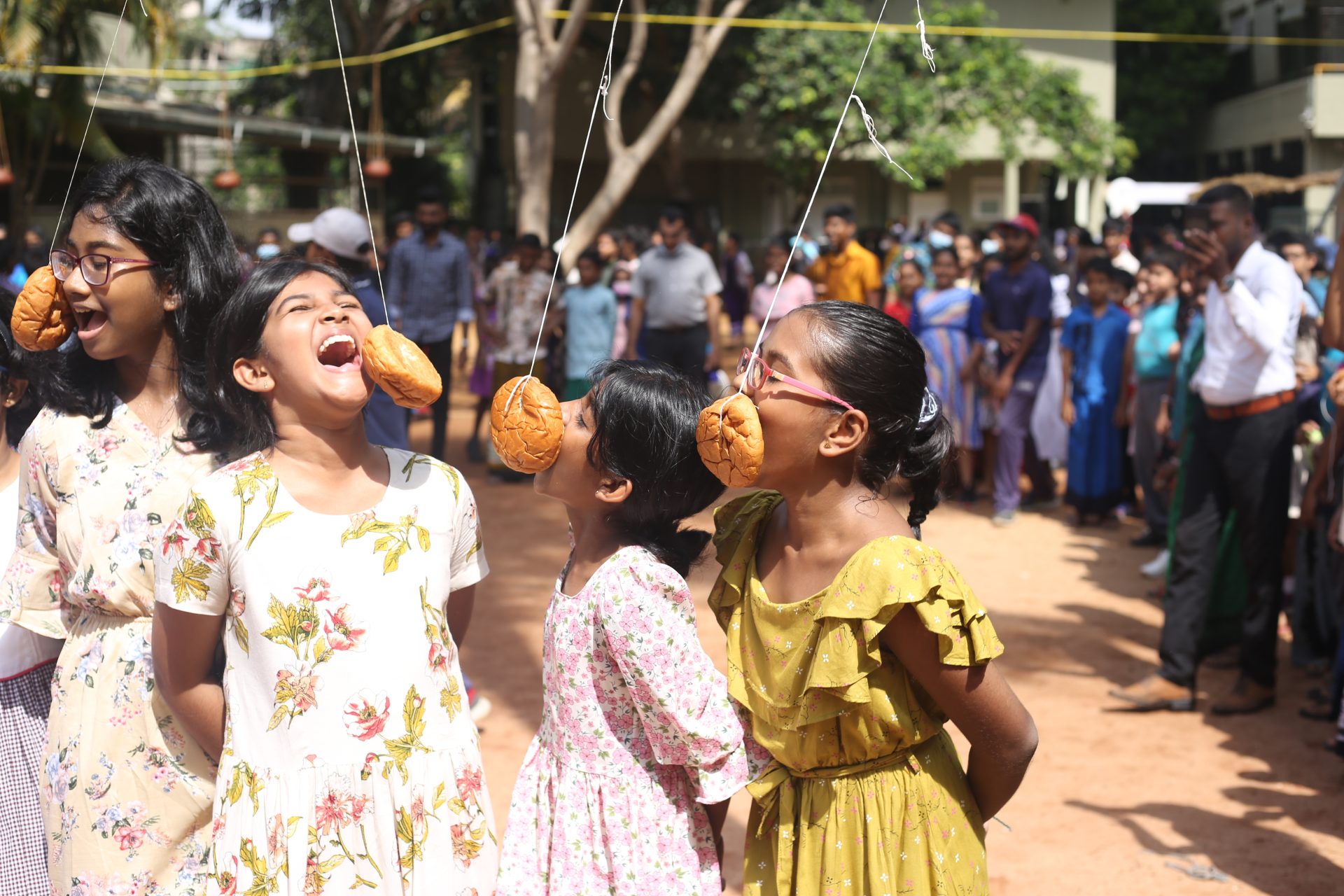 Children enjoy doughnut-eating contest at outdoor festival, surrounded by amused spectators.