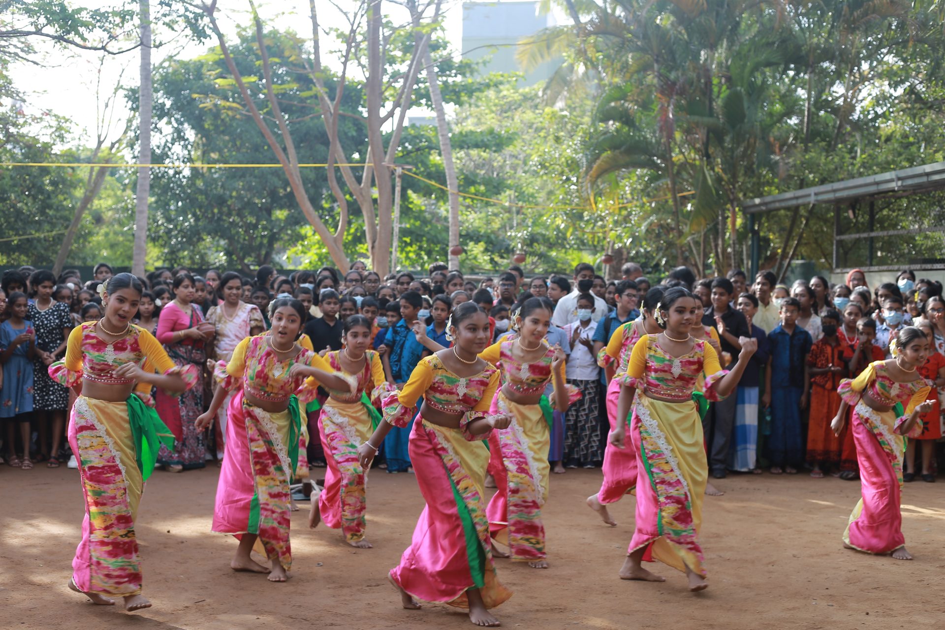 Young women perform traditional dance in colorful saris; a diverse crowd watches attentively.