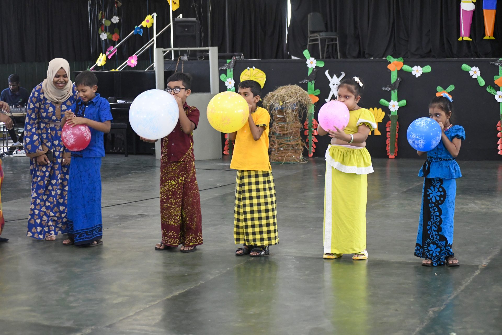 Kids in traditional attire enjoy a balloon activity with a supervising adult in a decorated gym.