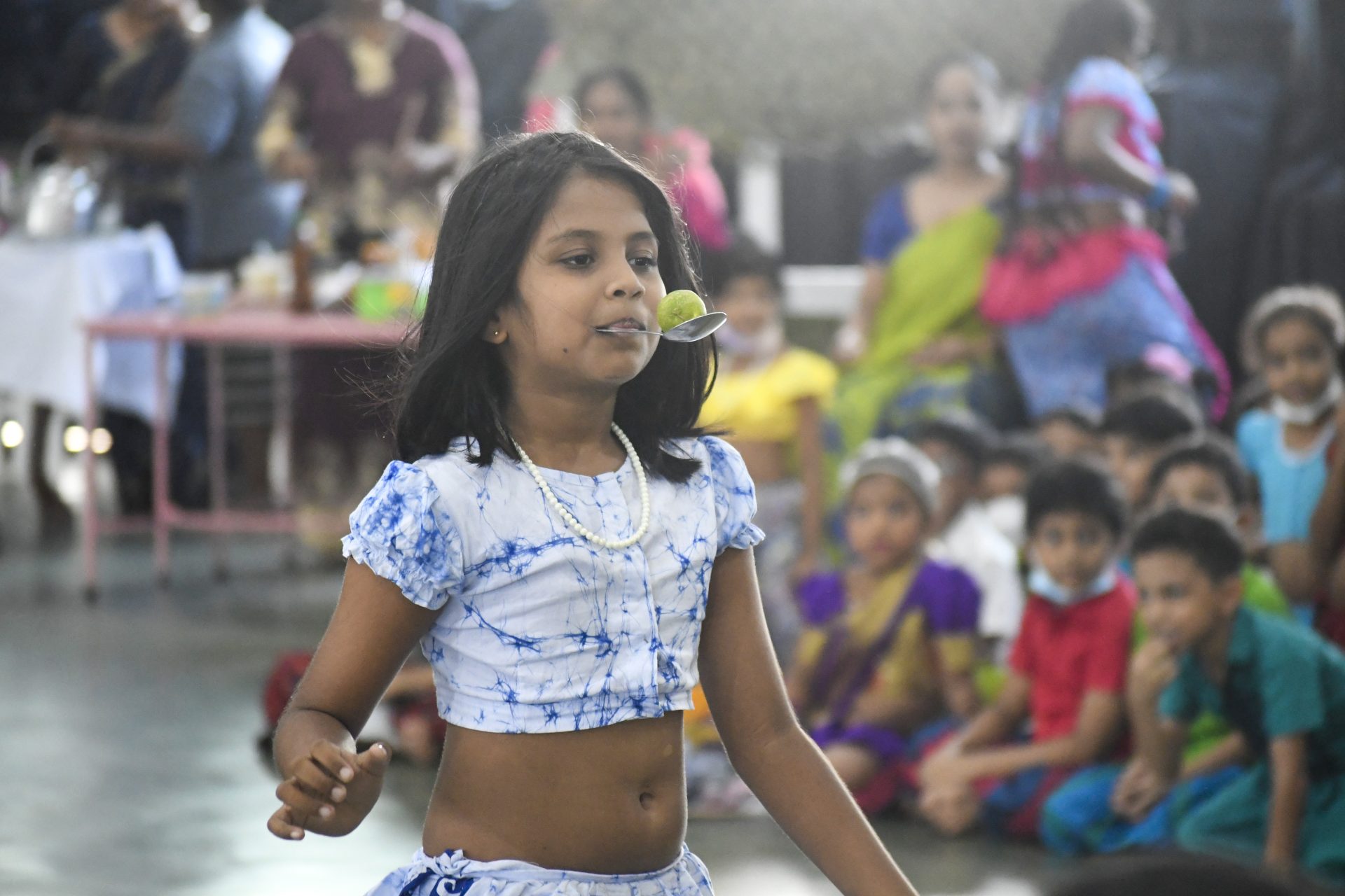 Girl in blue participates in lime-on-spoon game at vibrant indoor cultural event.