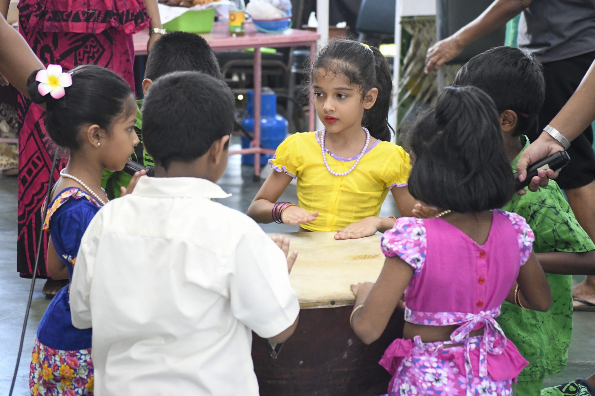 Children in colorful outfits playing a traditional drum indoors, celebrating a cultural event.