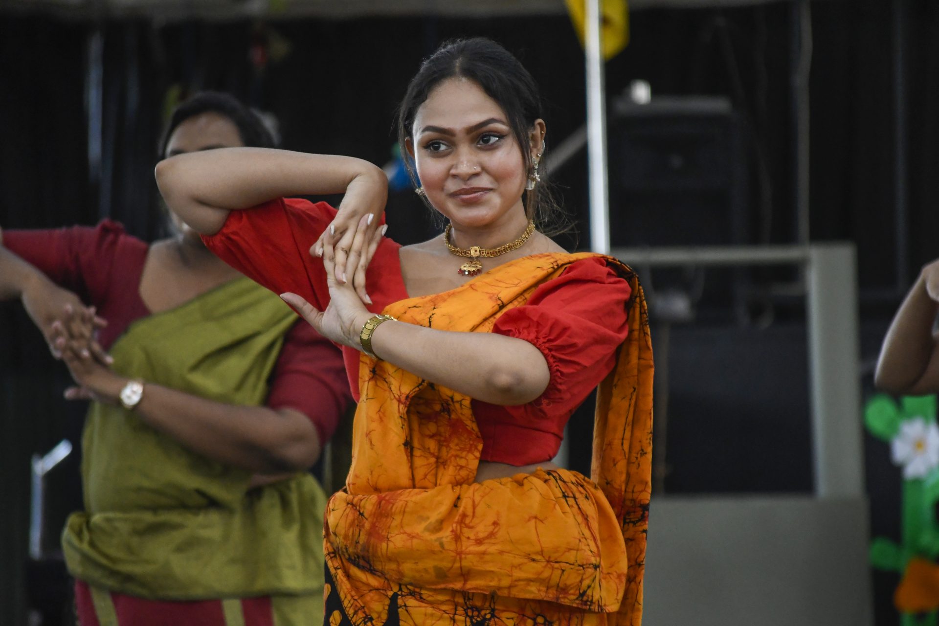 Woman in orange saree performs traditional dance; another dancer in green seen in the background.