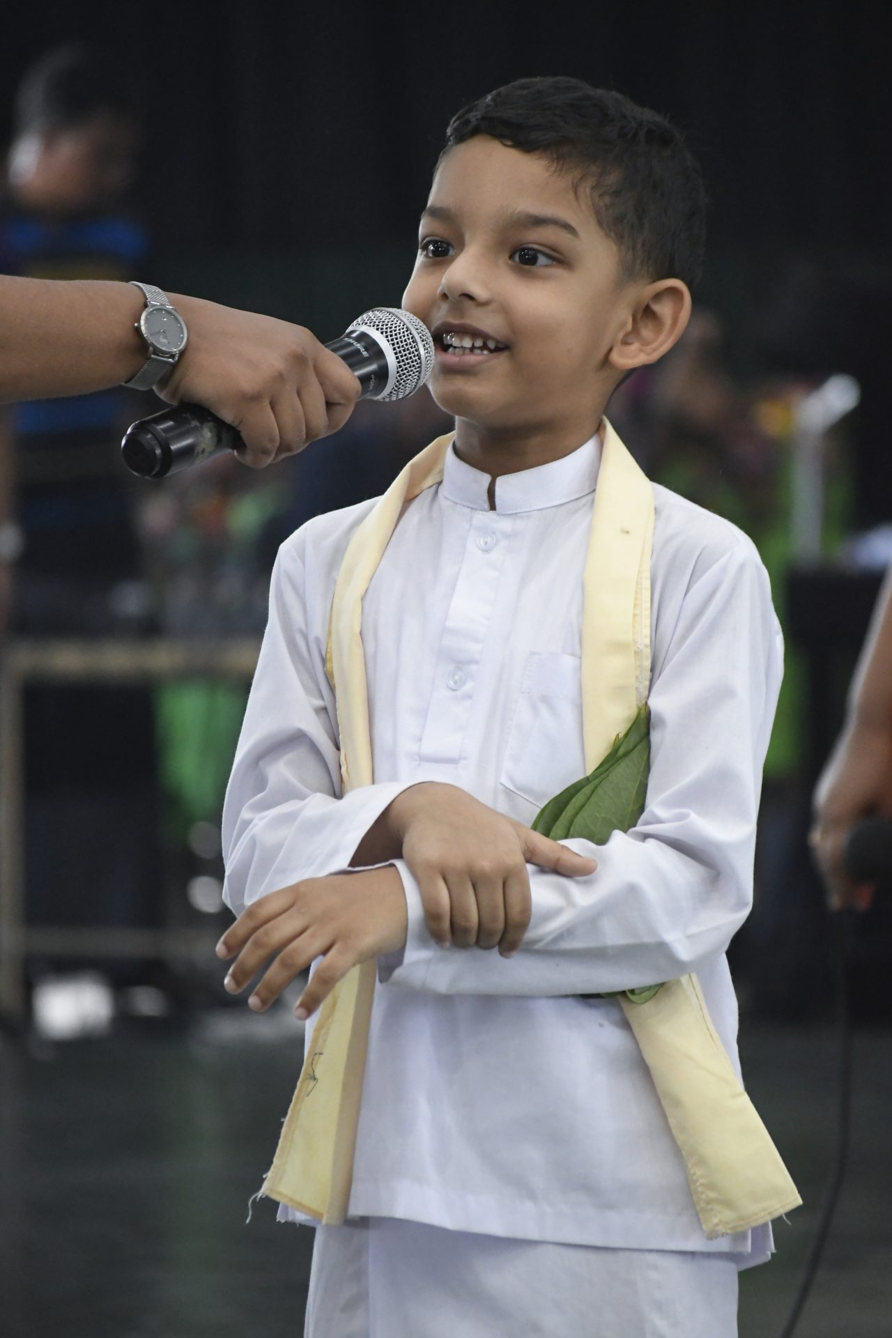 Young boy in traditional attire speaking at a cultural event.