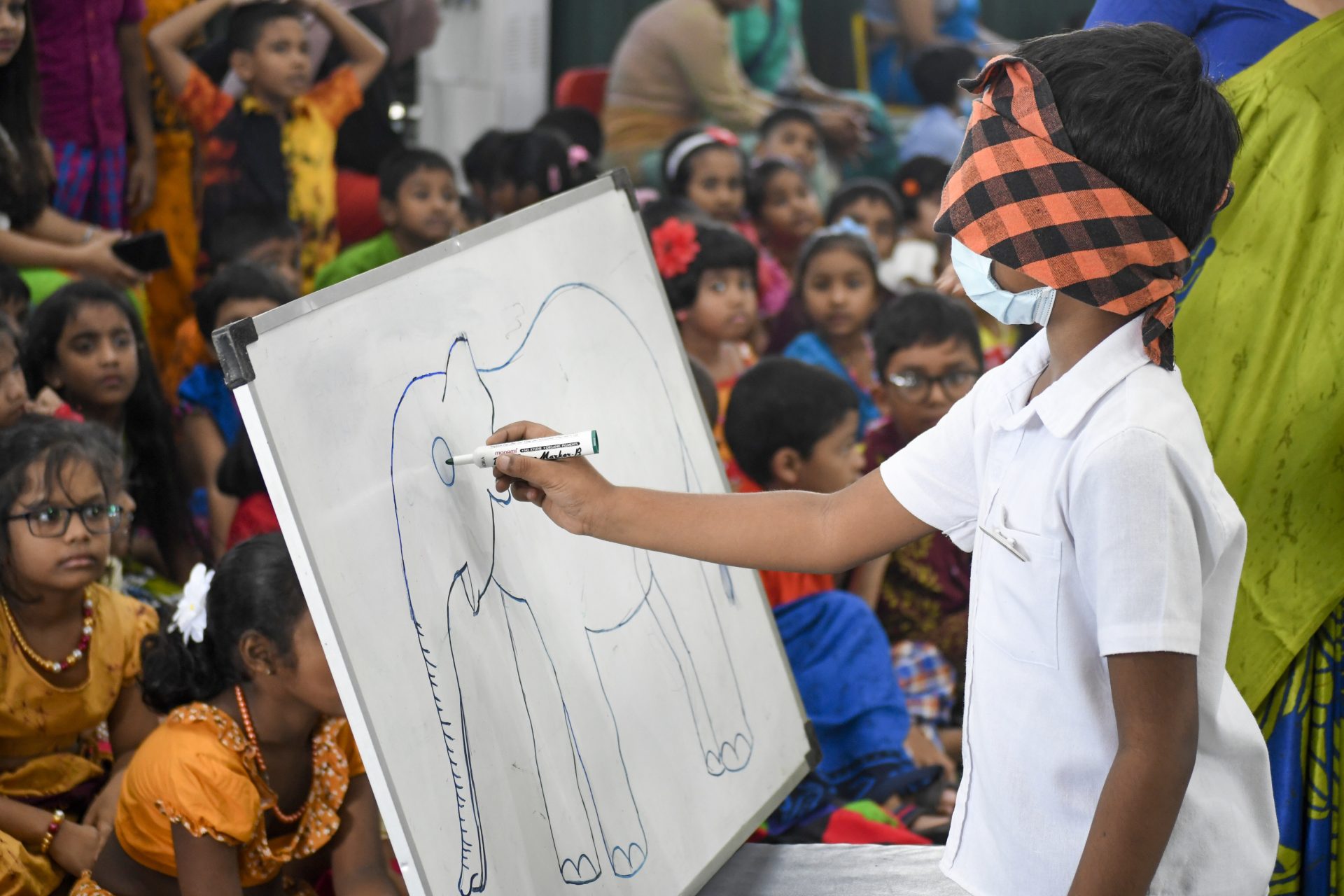 Blindfolded boy drawing elephant on whiteboard surrounded by children in traditional attire.