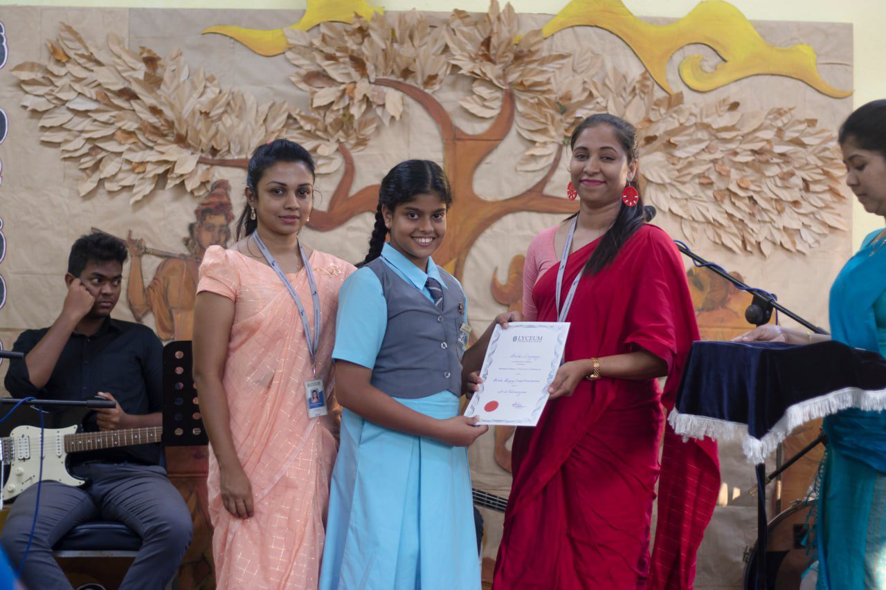 School award ceremony with student receiving certificate, musicians performing, and symbolic tree backdrop.
