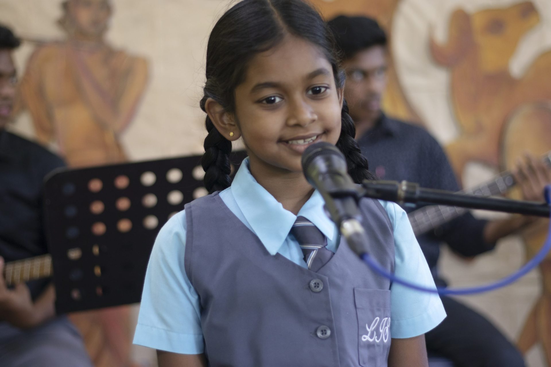 Schoolgirl sings confidently with musicians in front of a colorful mural.