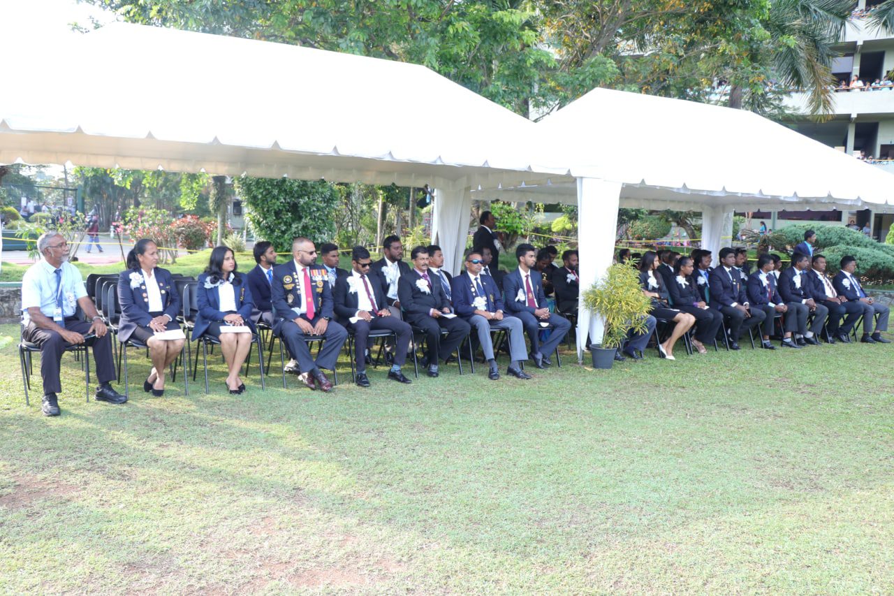 Outdoor graduation ceremony with attendees in formal attire under white canopies on a sunny lawn.