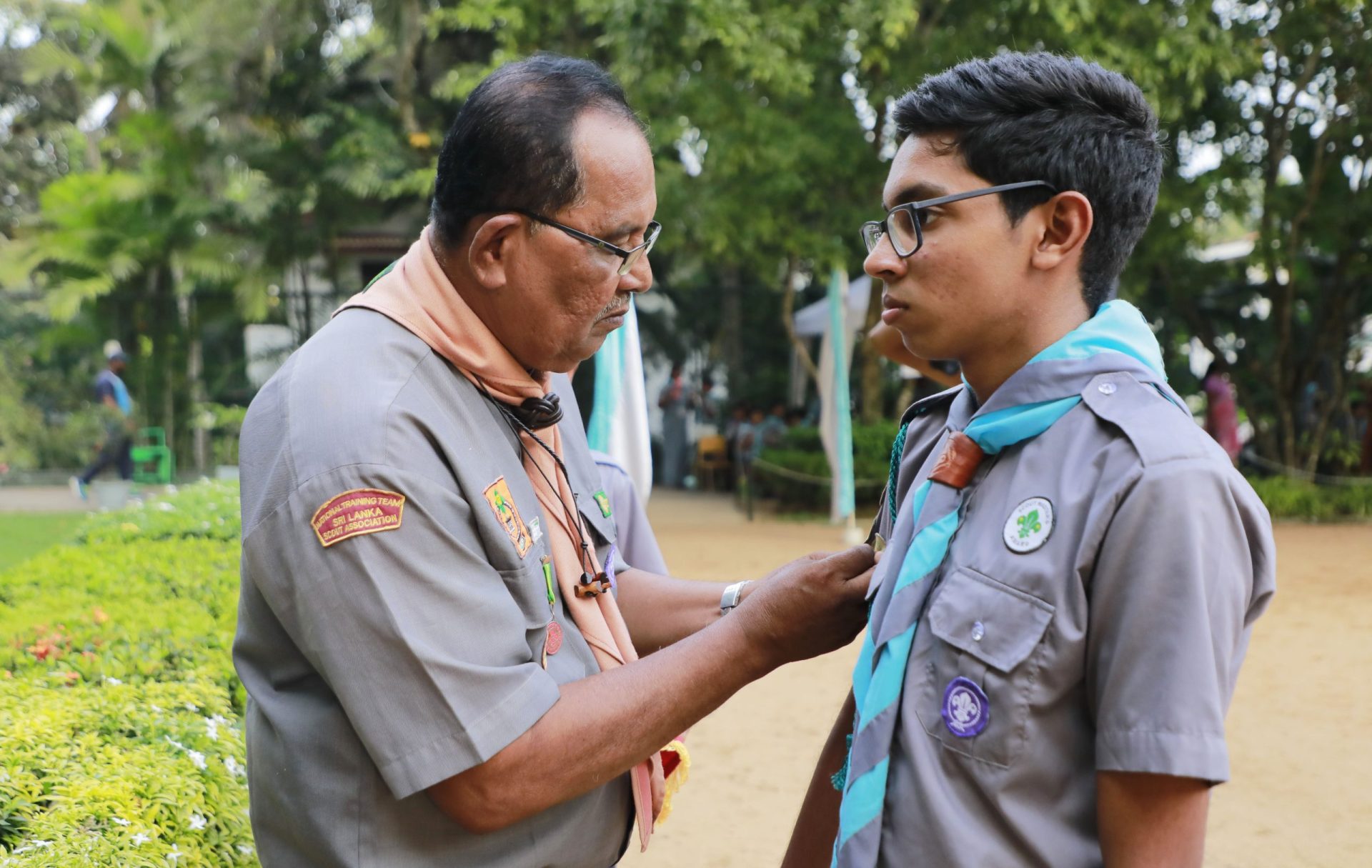 Scout leader pins badge on scouts uniform during outdoor ceremony in a lush park.
