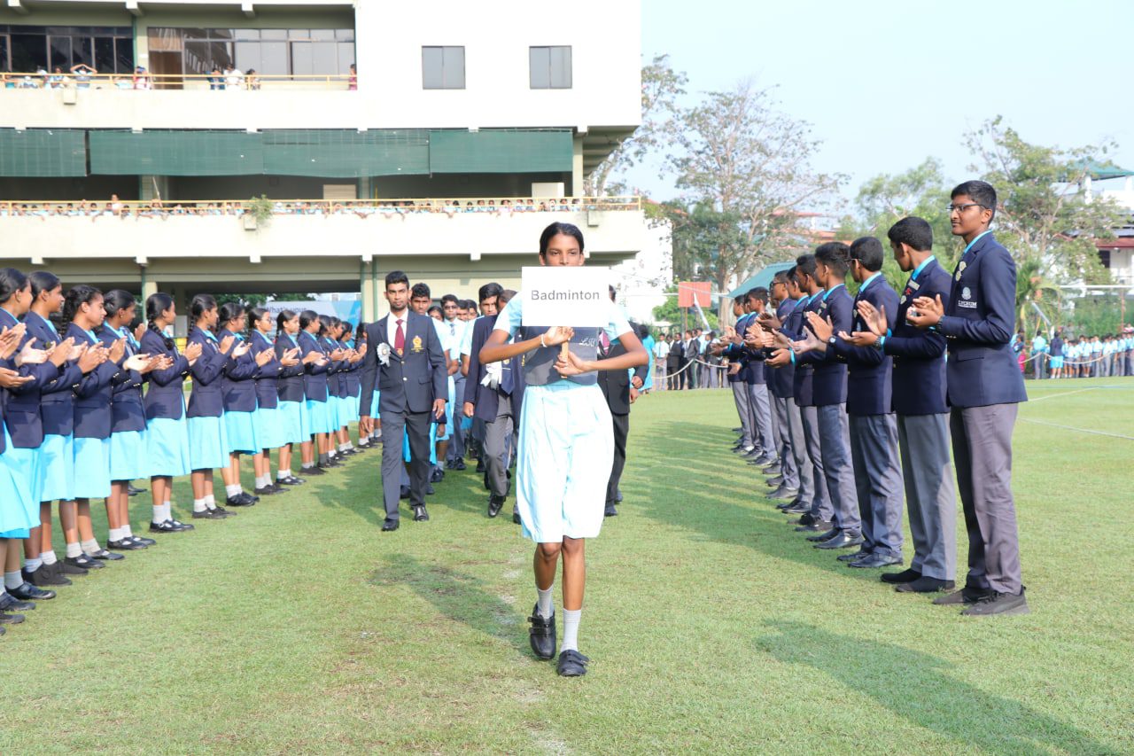 School sports day parade with students in uniforms and a girl leading with a placard.