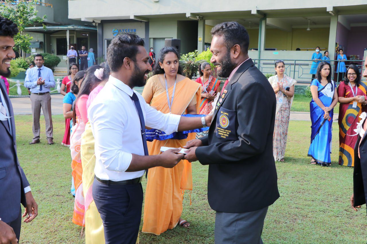 Award ceremony at Lyceum with men exchanging award, women in colorful sarees, modern building background.