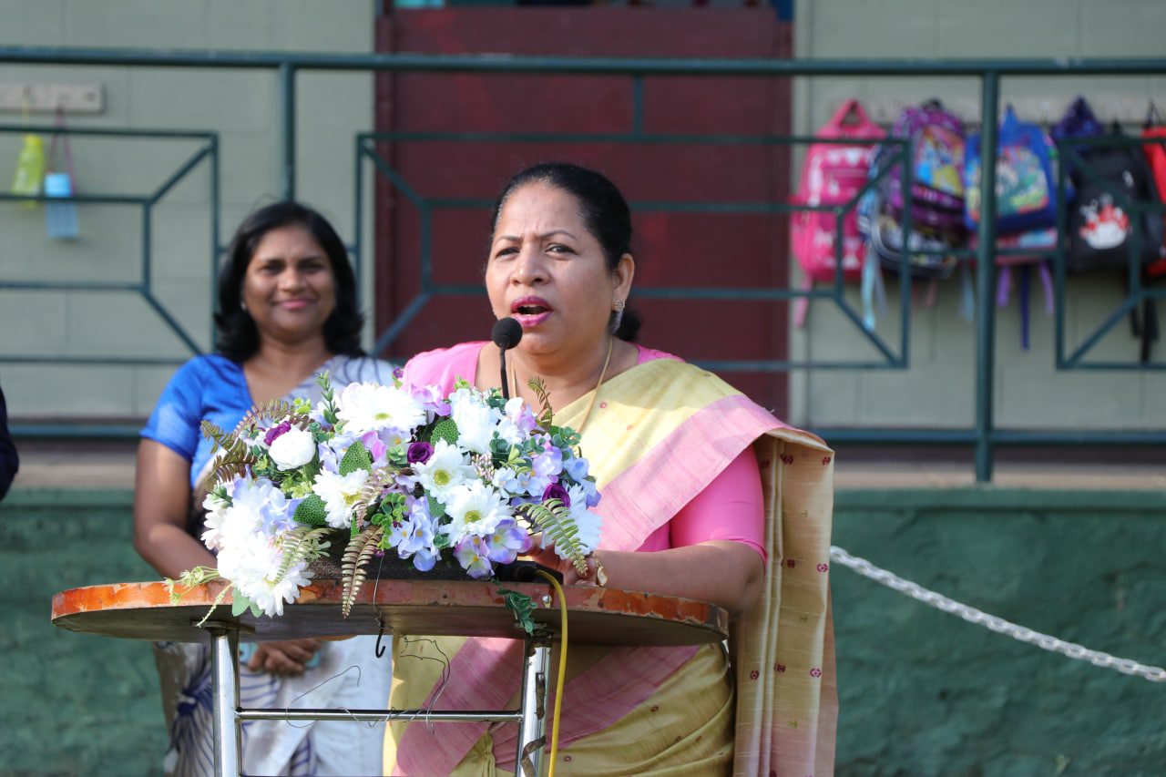 Woman in sari speaks at flower-adorned podium during school event.