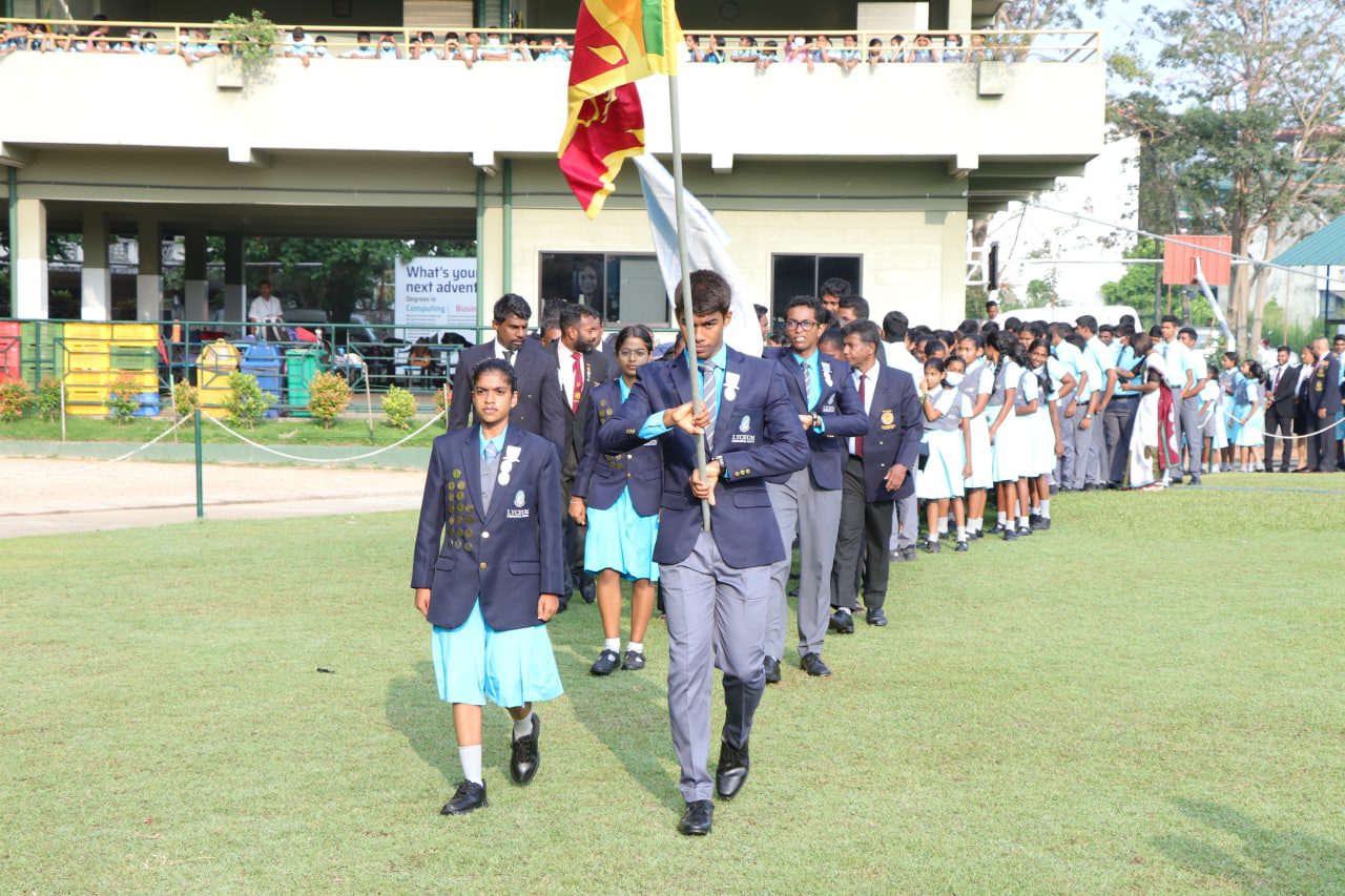 Students in uniform marching with a flag during a formal school ceremony.
