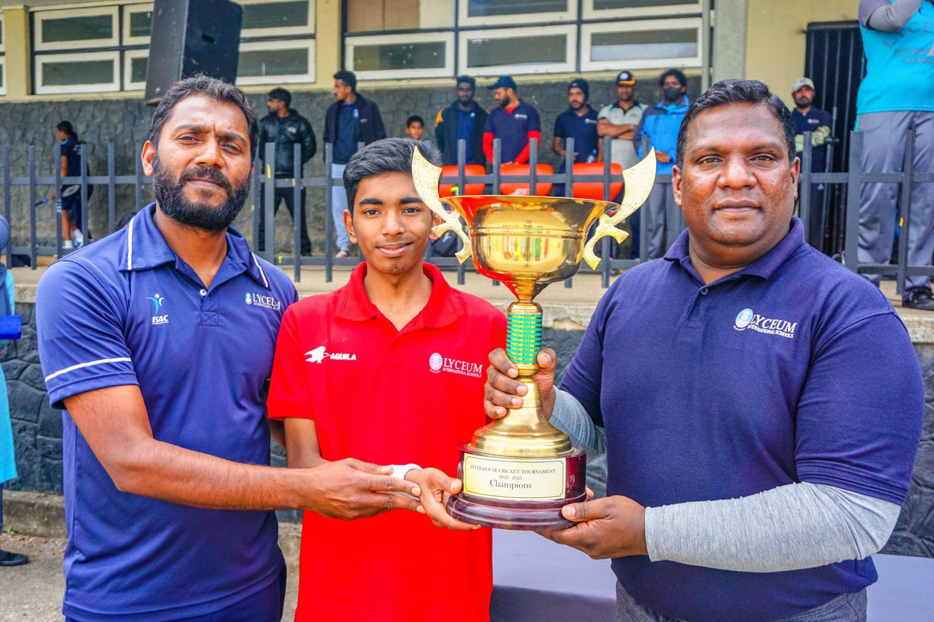 Three men celebrating with a large trophy at an outdoor sports event.