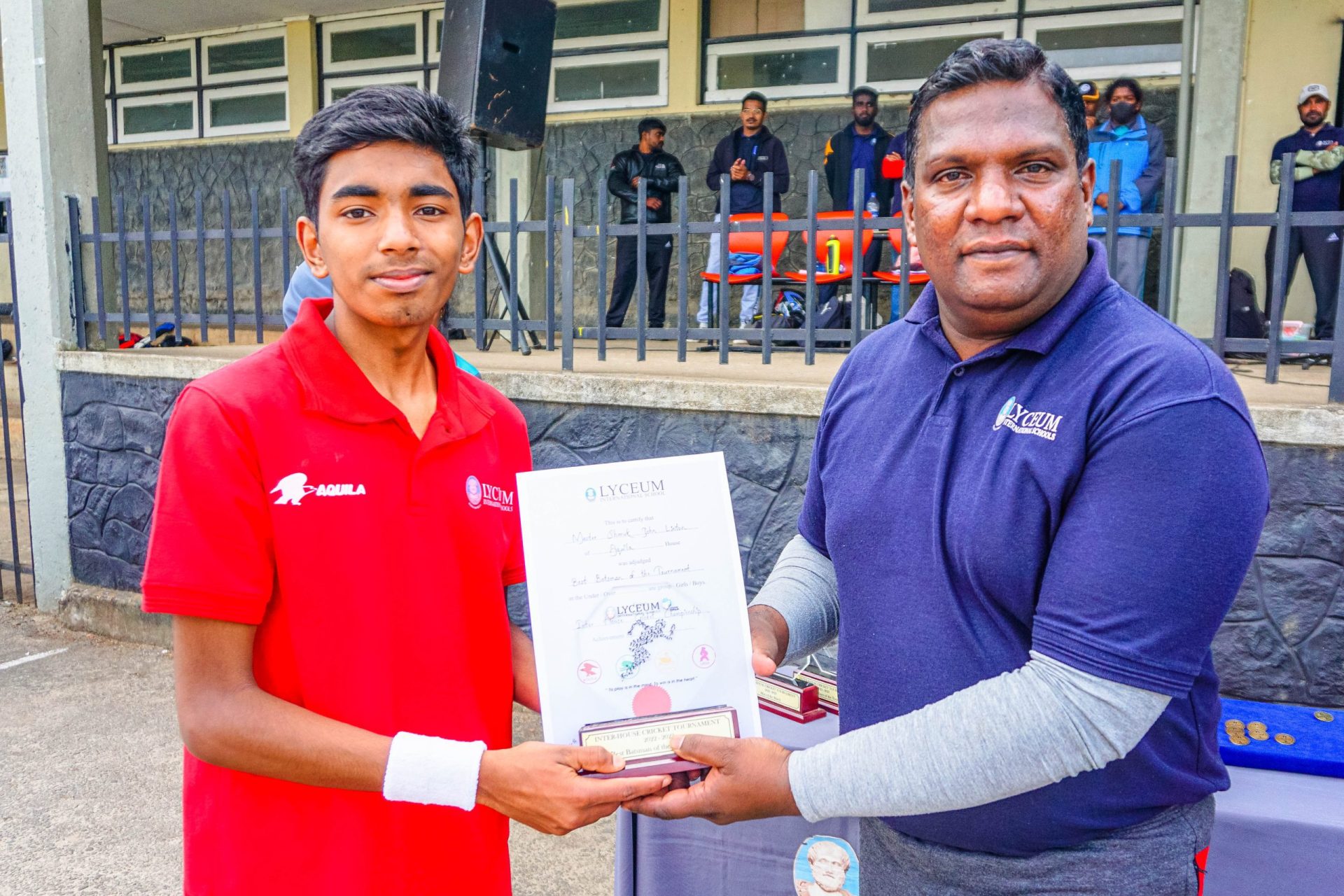 Young man in red shirt receives certificate at outdoor ceremony.