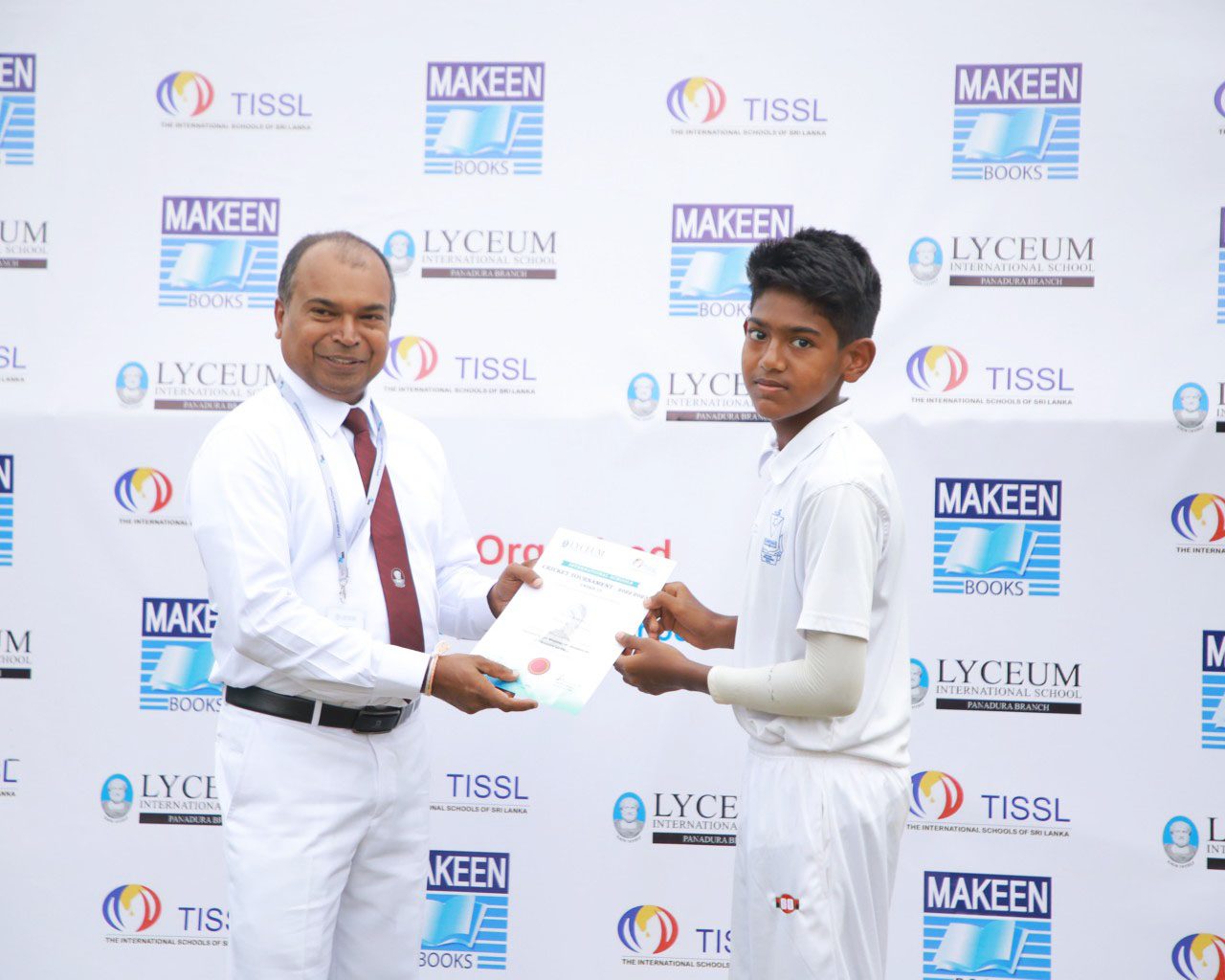 Man presenting certificate to student in white cricket uniform at award ceremony.