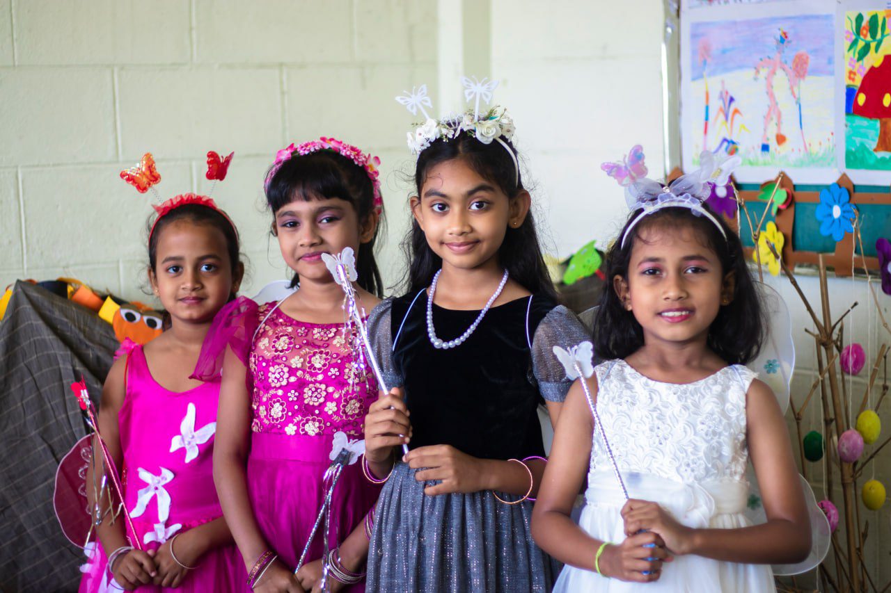 Four young girls in fairy costumes, holding butterfly wands in a decorated, festive setting.
