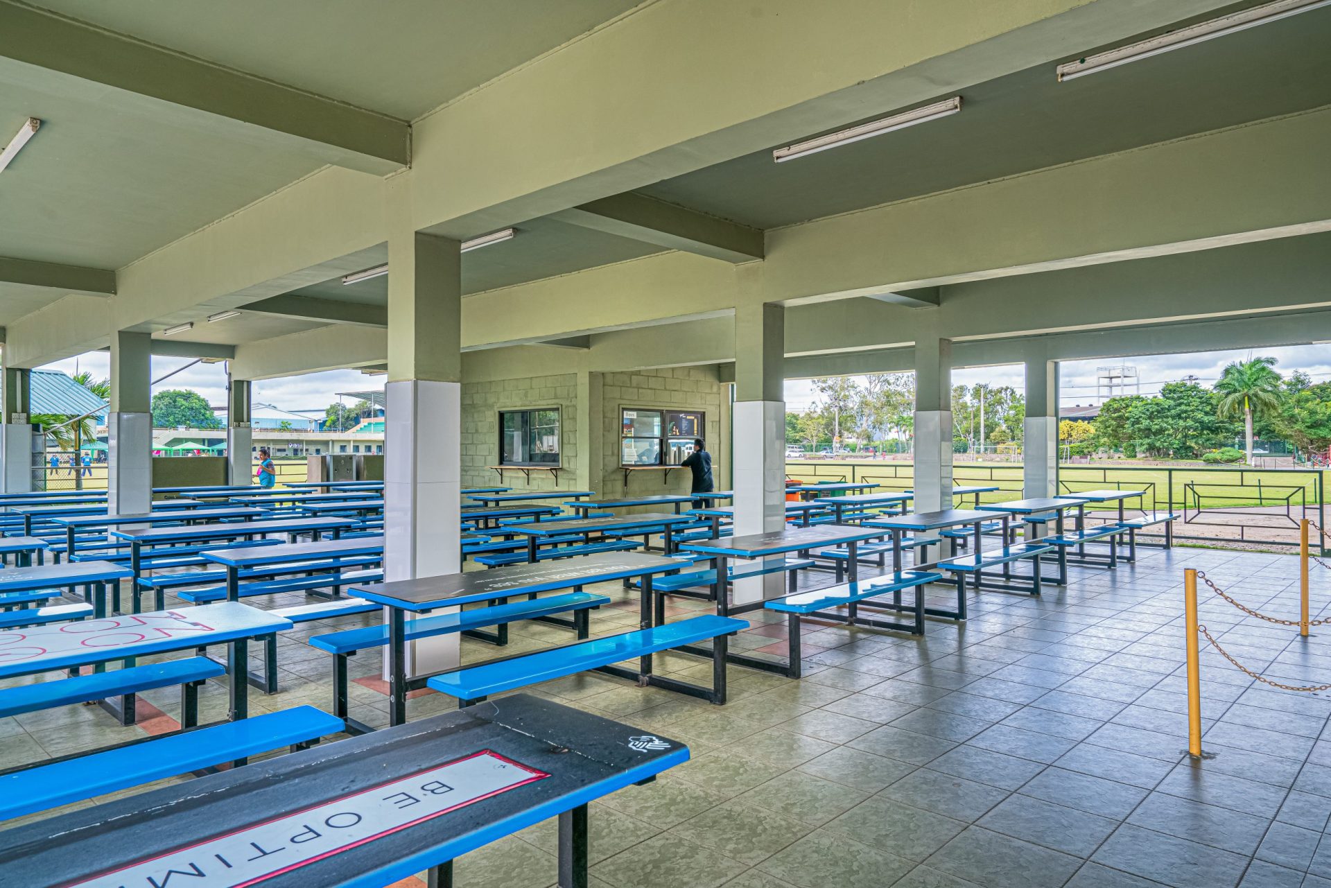 Open-air school cafeteria with blue tables, benches, and adjacent grassy field.