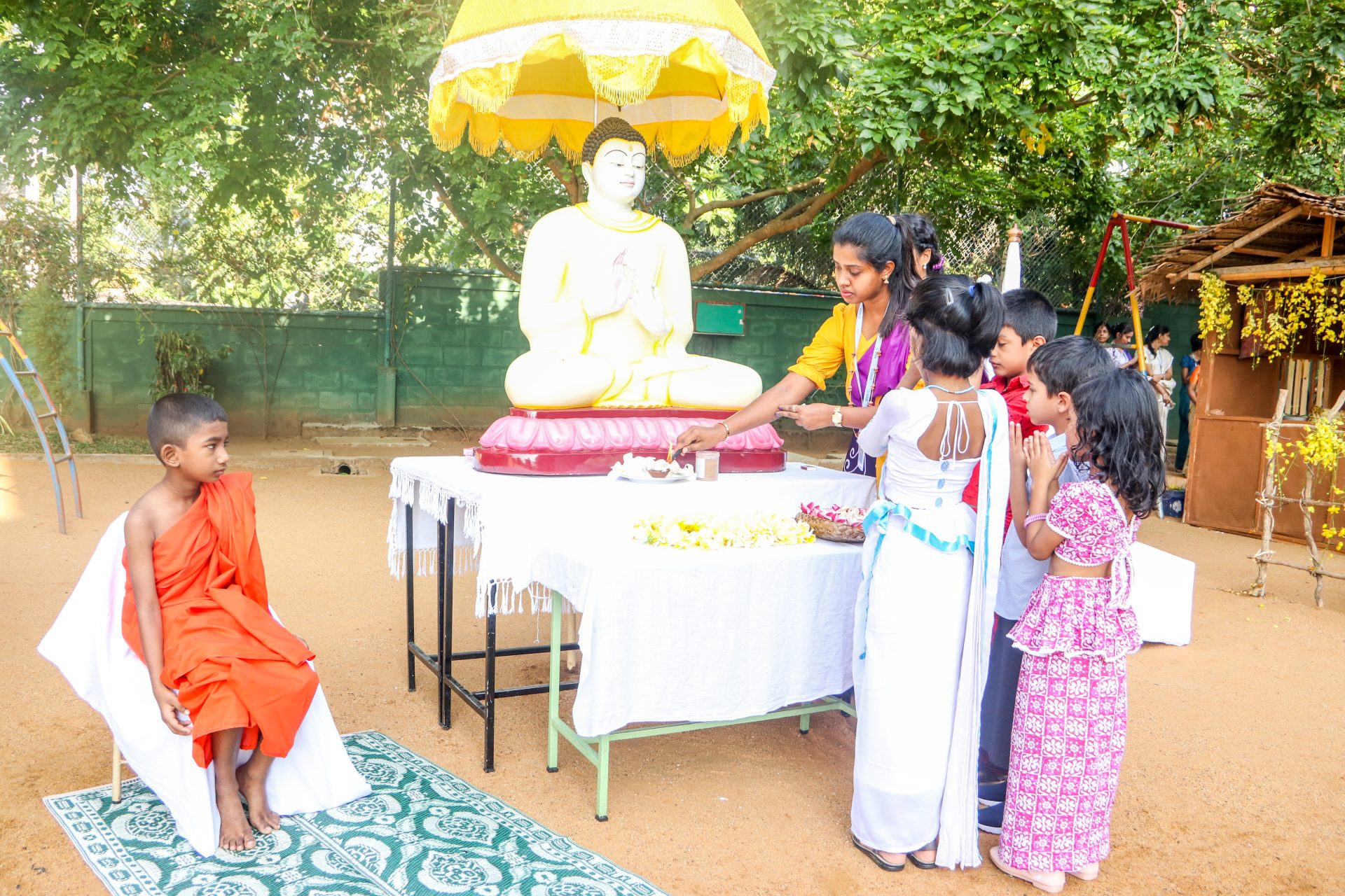 Children and monk honoring Buddha with offerings in a tranquil, lush outdoor ceremony.